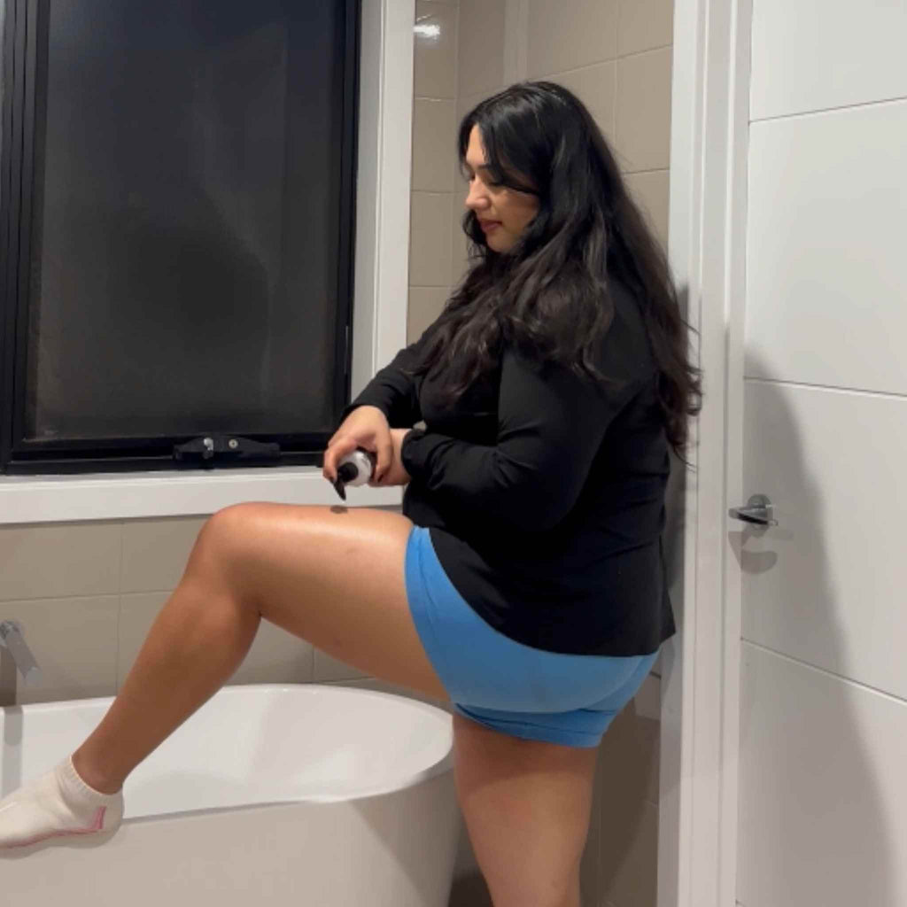 Woman sitting on a toilet in a bathroom applying lotion to her leg.