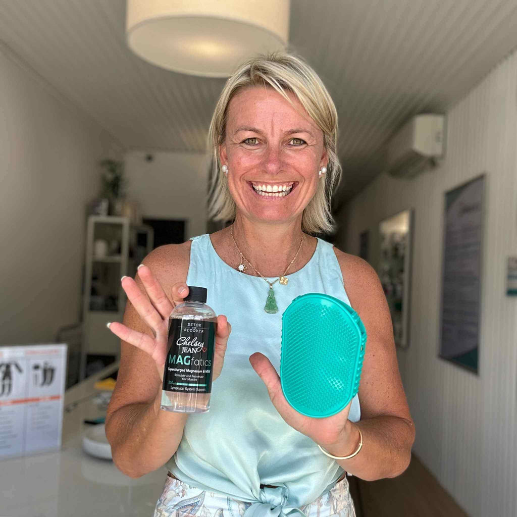 Chelsey holding the dual action lymphatic glove and the 250ml magfatics oil in a room with a white wall and ceiling light.